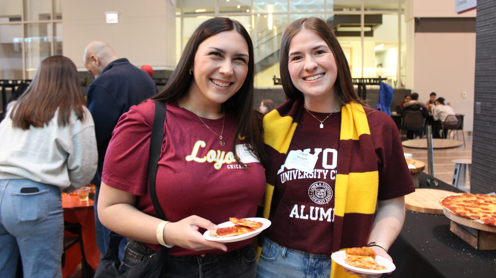 Two alumnae in Loyola maroon tee shirts and gear have pizza at a Loyola event